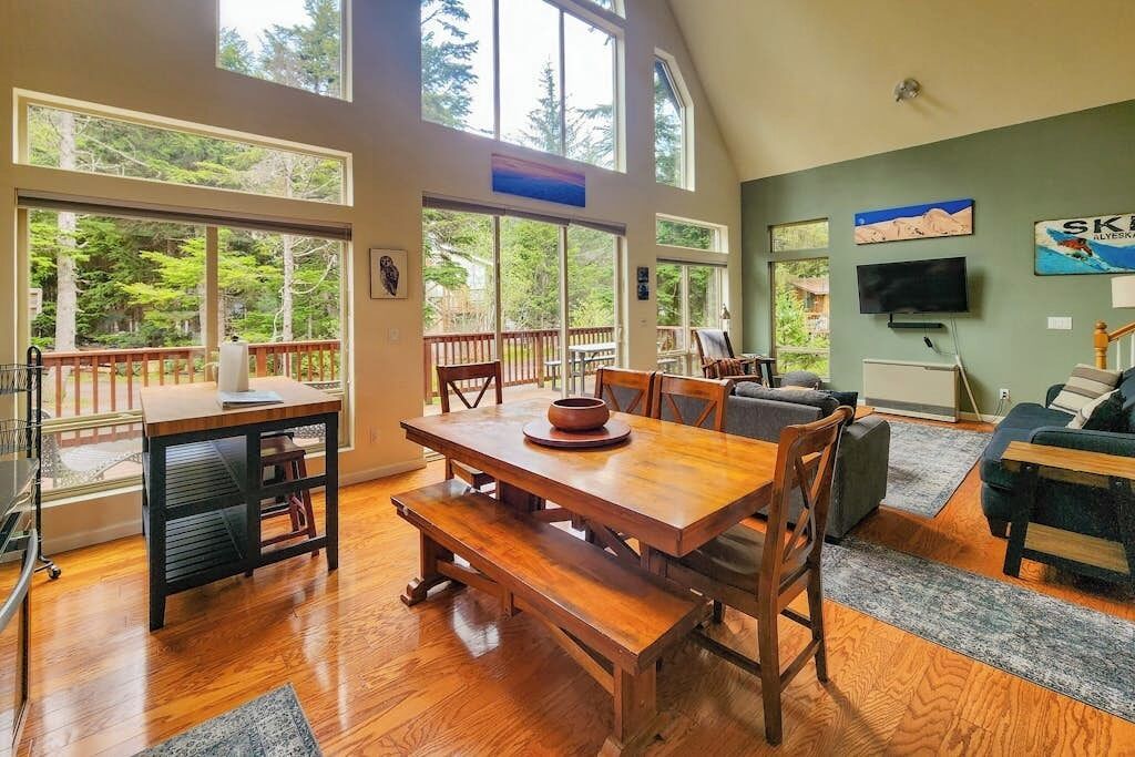A spacious living area with a wooden dining table, comfortable seating, and large windows offering a view of the outdoors.