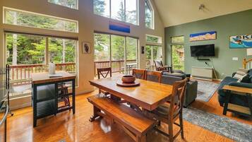 A spacious living area with a wooden dining table, comfortable seating, and large windows offering a view of the outdoors.