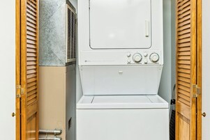 Pequeña lavandería blanca con lavadora y secadora integradas, puertas de madera con persianas y pared con azulejos.