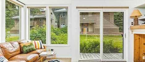 Un salon moderne avec un canapé en cuir, une petite table ronde et une vue sur une terrasse en bois, accessible par de grandes portes coulissantes en verre.