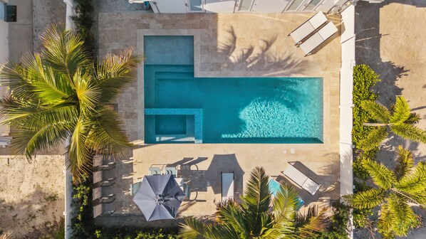 Vue aérienne d’une piscine au design géométrique, entourée de palmiers et de chaises longues.