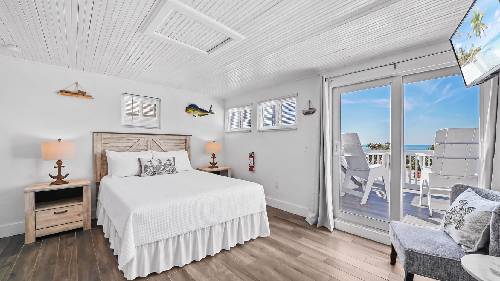 A bedroom with a large bed, a nightstand, a lamp, a window with blinds, and a view of the ocean through a sliding door.