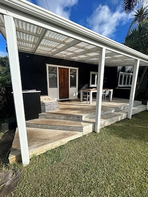 A covered outdoor area with a wooden deck, a door, and a table with chairs. - Oneroa Holiday House (Waiheke Island)