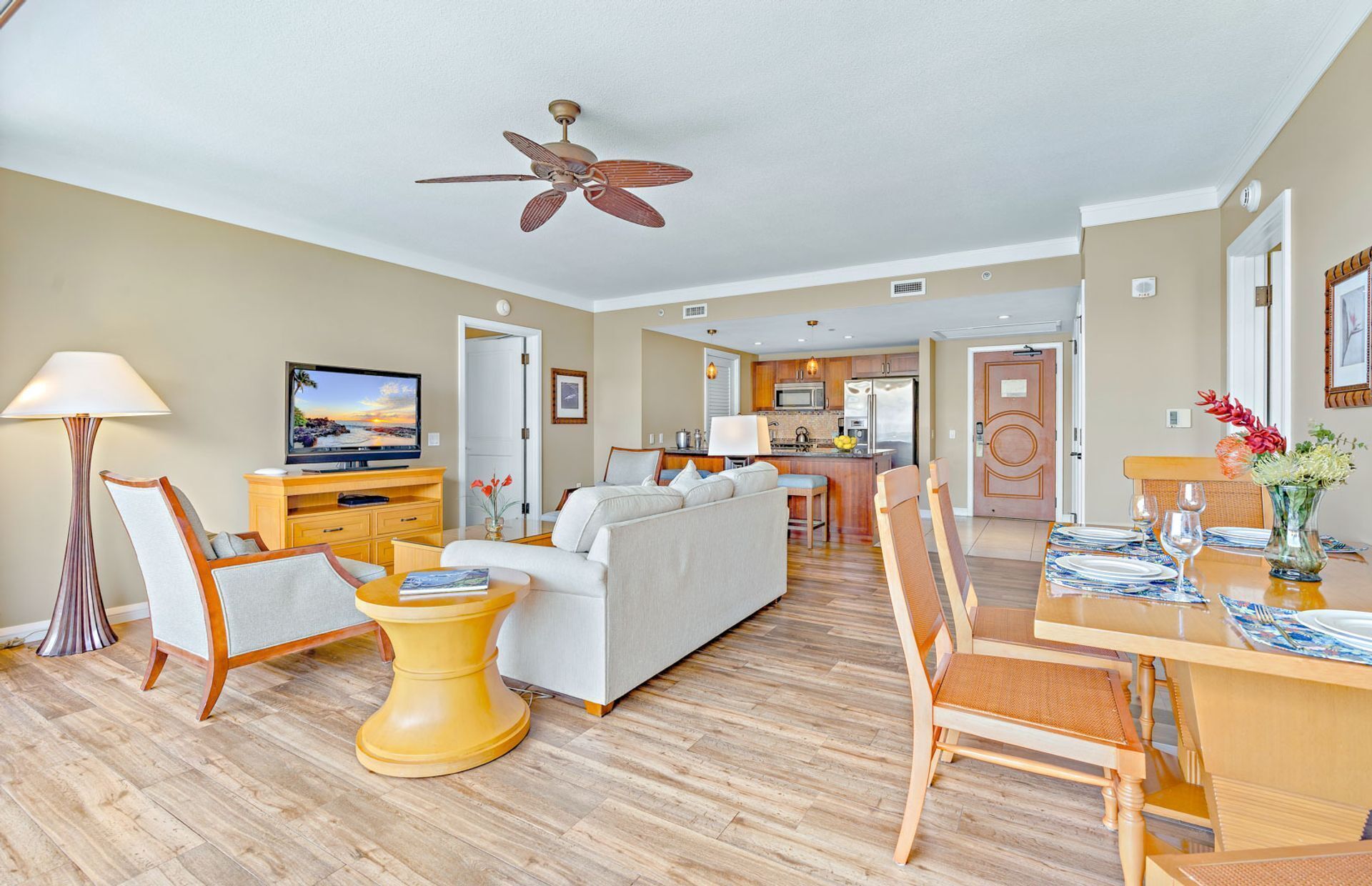 A modern living room with a sofa, a coffee table, and a television. There is a dining area with a table set for two. The room has wooden flooring and a ceiling fan.