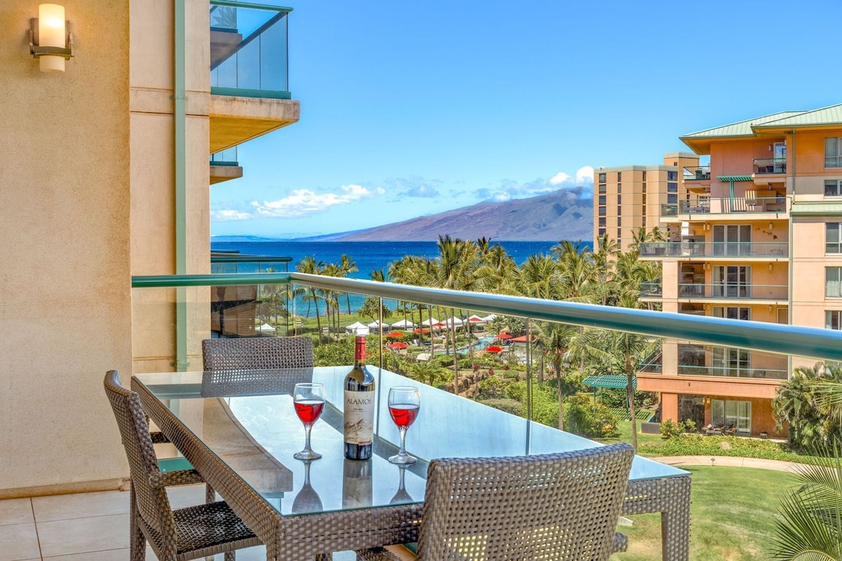 Ein Balkon mit Tisch, zwei Stühlen und zwei Weingläsern mit Flaschen, der Blick auf die Küste mit Gebäuden und einem Berg freigibt.