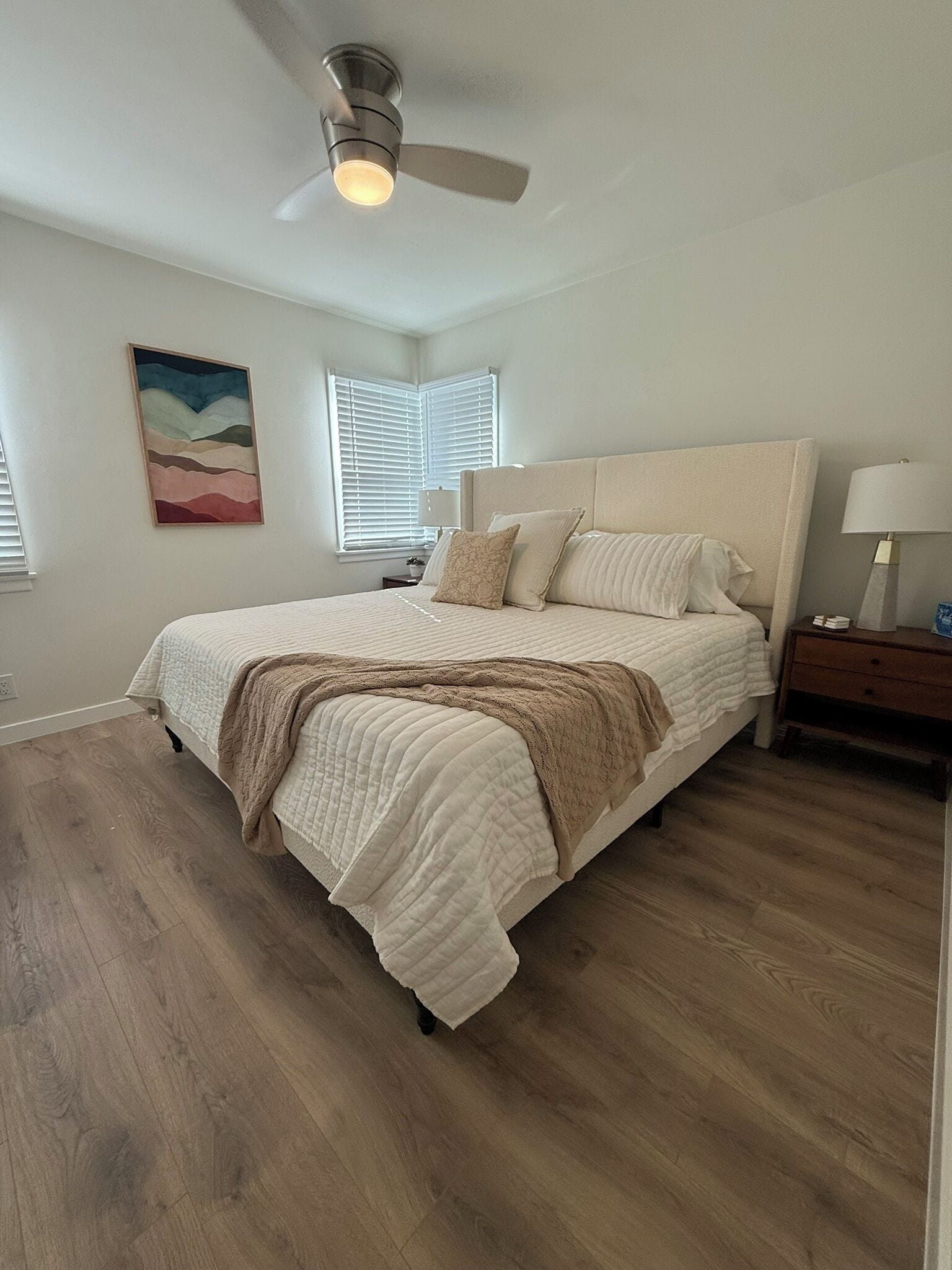 A bedroom with a bed, a nightstand, a lamp, a ceiling fan, and a painting on the wall.