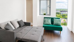 A modern living room with a grey sofa, a green armchair, and a large window offering a city view. - Modern 1BR Flat Sleeps 5 in Wimbledon (London)