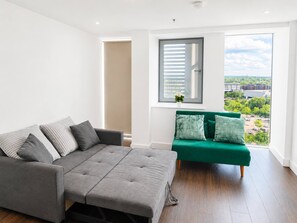 Ein modernes Wohnzimmer mit grauem Sofa, einem grünen Sessel und einem großen Fenster mit Blick auf die Stadt.
