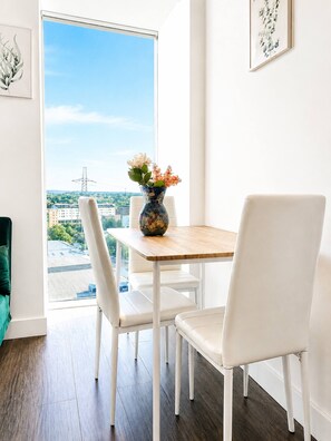 Ein Essbereich mit Tisch und Stühlen, einer Blumenvase und Blick auf die Stadt durch ein großes Fenster.