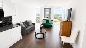 A modern living space with a kitchen, a grey sofa, a green sofa, a round coffee table, a white chair, and a TV on a wooden stand. - Modern 1BR Flat Sleeps 5 in Wimbledon (London)