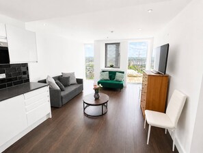 Ein zeitgemäßer Wohnraum mit Küche, einem grauen Sofa, einem grünen Sofa, einem runden Couchtisch, einem weißen Sessel und einem Fernseher auf einem Holzgestell.