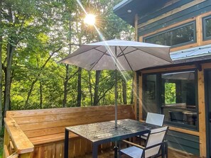 Eine Holzterrasse mit Tisch und Stühlen, einem Sonnenschirm und Blick auf ein bewaldetes Gebiet.