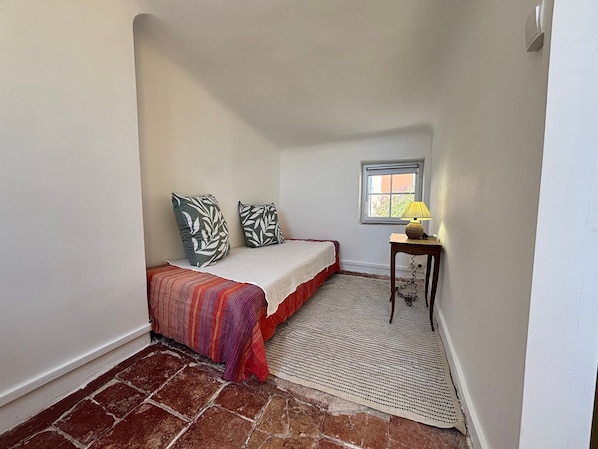 A small bedroom with a single bed, a window, a table with a lamp, and a patterned rug. - Au Levant, Charming Village House with Sea View (Bormes-les-Mimosas)