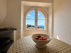A room with a round table, a bowl of fruit, a window with a view of buildings, and a shelf with items. - Au Levant, Charming Village House with Sea View (Bormes-les-Mimosas)