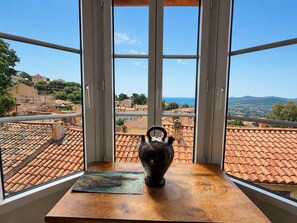A room with a view of a coastal town through large windows, a wooden table with a vase and a painting, and a tiled roof visible outside. - Au Levant, Charming Village House with Sea View (Bormes-les-Mimosas)