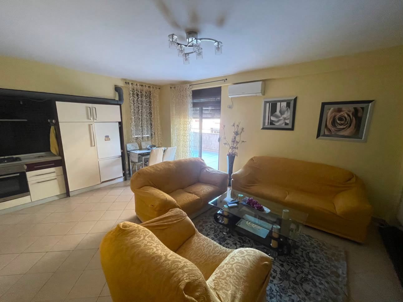 Sala de estar com sofá, mesa de centro e televisão. Há uma cozinha com armários e geladeira.