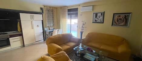 A living room with a sofa, a coffee table, and a TV. There is a kitchen area with cabinets and a refrigerator.
