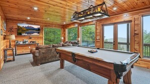 A wooden interior with a pool table, a sofa, and a large window offering a view of the outdoors. - High Smokies Lodge - Game Room, Hot Tub and more! (Sevierville)