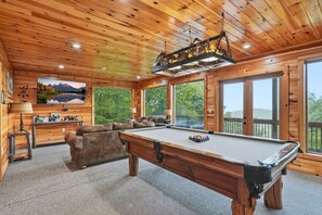 A wooden interior with a pool table, a sofa, and a large window offering a view of the outdoors. - High Smokies Lodge - Game Room, Hot Tub and more! (Sevierville)