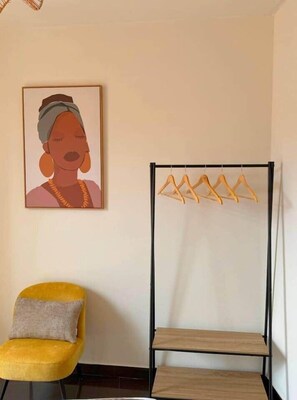 Ein Zimmer mit einem gelben Sessel, einem gerahmten Kunstwerk und einem schwarzen, metallenen Wäscheständer mit Holzaufhängern.