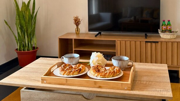 A modern living room with a wooden coffee table, two plates of pastries, a glass of milk, a television, a potted plant, and a small basket of snacks. - Church View by Stayguest (Santa Cruz)