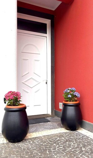 Eine Tür mit geometrischem Muster, eingerahmt von zwei großen, schwarzen Pflanzgefäßen mit Blumen, vor einer roten Wand.