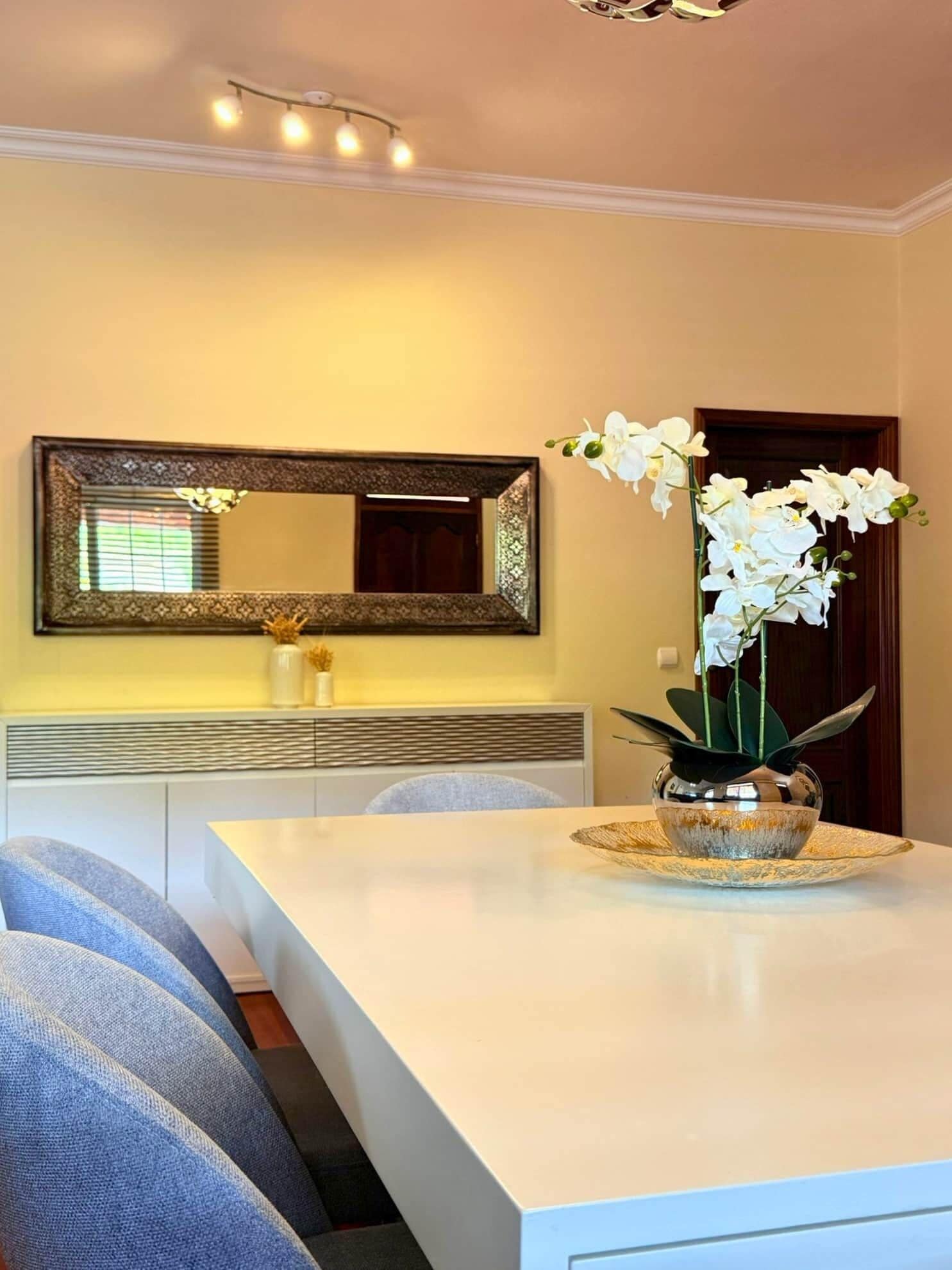 A dining area with a white table, a vase of white flowers, and a mirror above the counter.