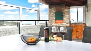 A table with a bottle of wine, two glasses, and a basket of oranges on a terrace with a view. - Yara House by Stayguest (Câmara De Lobos)