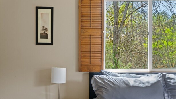 Ein Schlafzimmer mit Bett, Nachttisch mit Lampe, einem gerahmten Bild an der Wand und einem Fenster mit Blick auf Bäume.