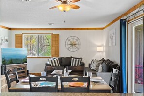 A cozy living room with a television, a dining table set for four, and a large window with a view of trees. - Mountain View Loft | Cozy Single-Level Retreat (Gatlinburg)