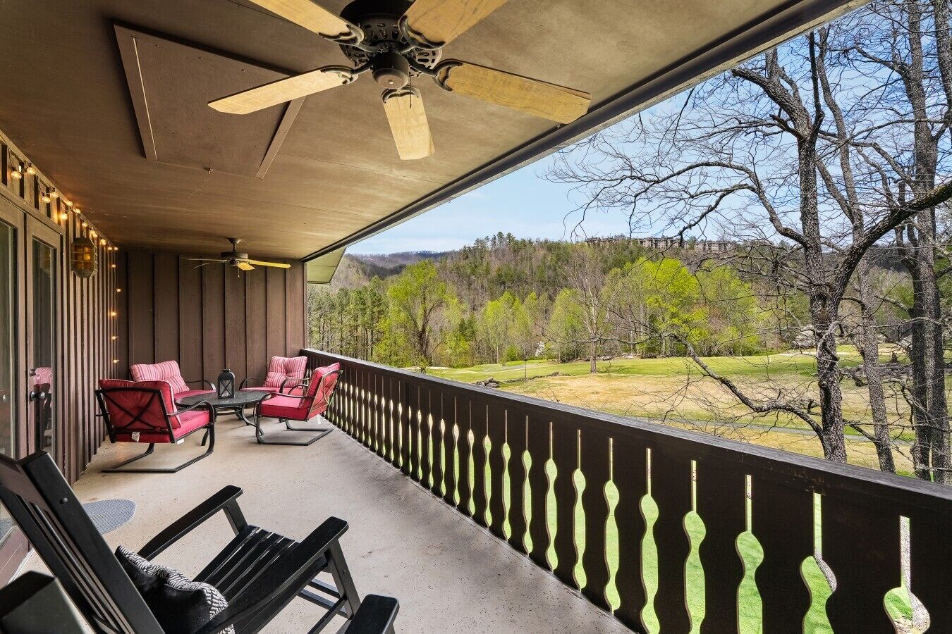 A covered porch with a ceiling fan, red cushioned chairs, and a view of a green landscape.