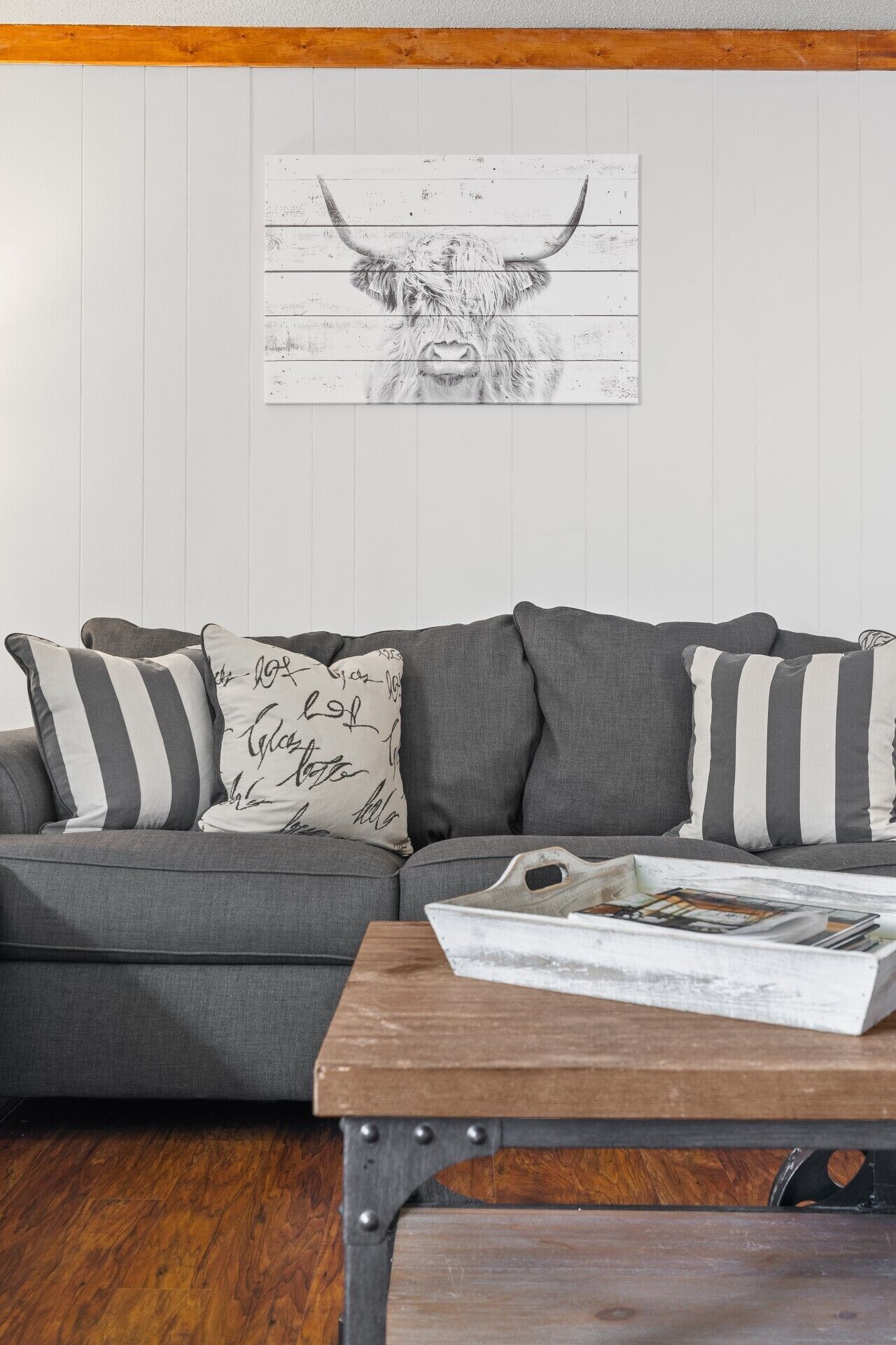 Een grijze bank met gestreepte kussens, een houten salontafel met tijdschriften, en een kunstwerk aan de muur van een stierenkop.