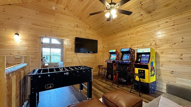 A room with a foosball table, two arcade machines, a television, and a ceiling fan. - Serene MTN Cabin: Indoor Pool, Hot Tub, & Game Room! (Pigeon Forge)
