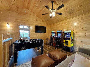 A room with a foosball table, two arcade machines, a television, and a ceiling fan. - Serene MTN Cabin: Indoor Pool, Hot Tub, & Game Room! (Pigeon Forge)