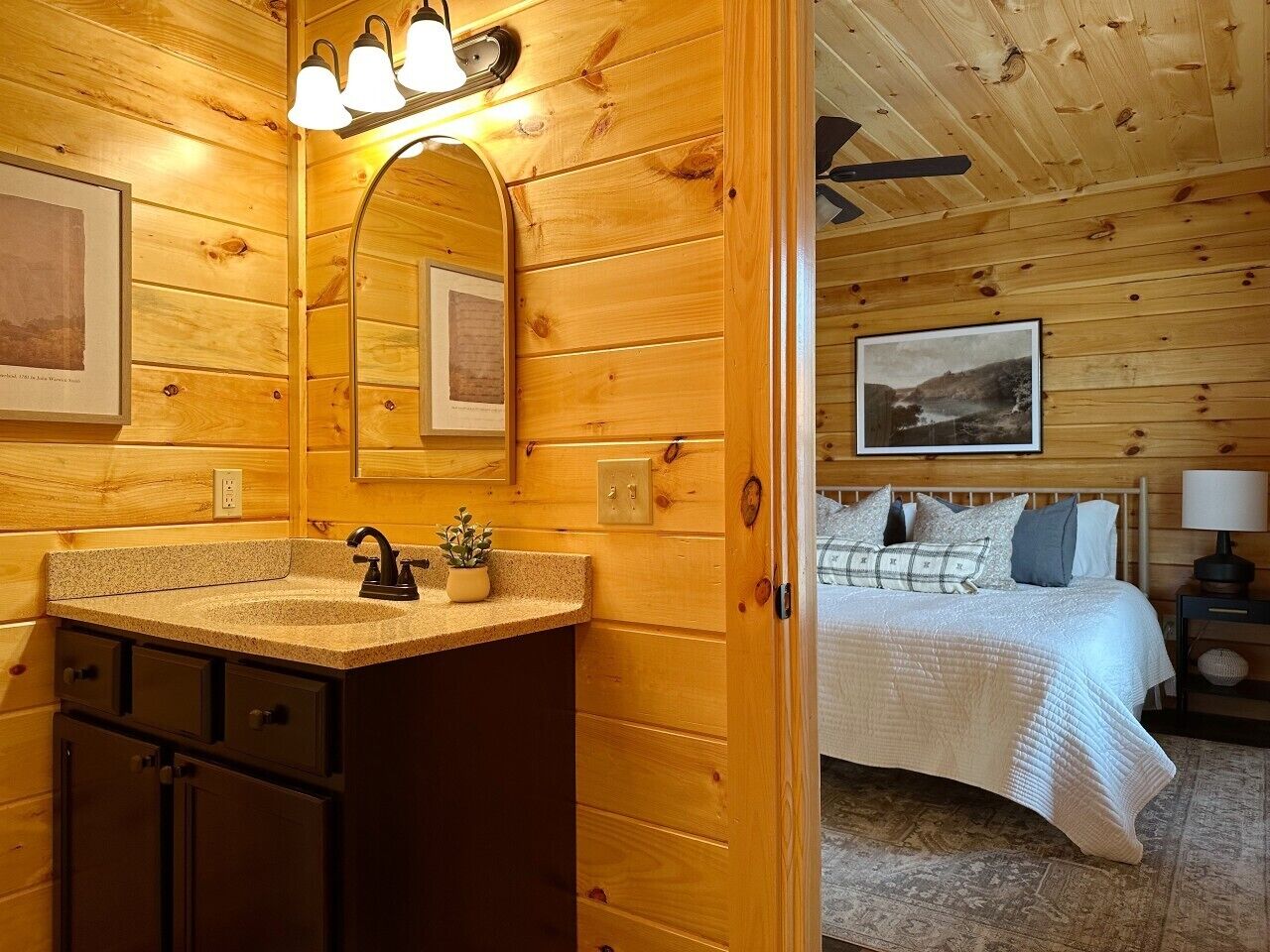 A wooden cabin interior with a bathroom featuring a sink, mirror, and vanity, and a bedroom with a bed and nightstand.