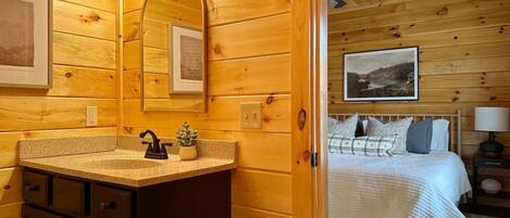 A wooden cabin interior with a bathroom featuring a sink, mirror, and vanity, and a bedroom with a bed and nightstand.