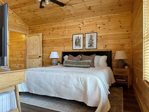 Quarto de cabana de madeira com uma cama, duas mesas de cabeceira com abajures, uma escrivaninha e uma janela com persianas.
