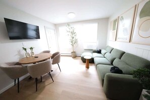 A modern living room with a flat-screen TV, a round wooden table with chairs, a green sofa, and a potted plant.
