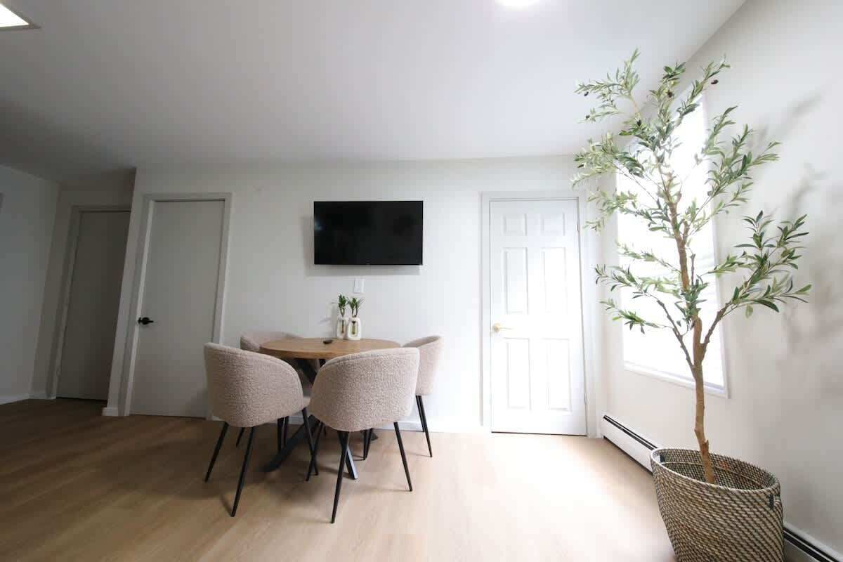 A room with a dining table, four chairs, a flat-screen TV mounted on the wall, and a potted plant.