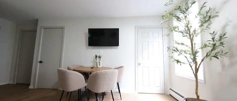 A room with a dining table, four chairs, a flat-screen TV mounted on the wall, and a potted plant.
