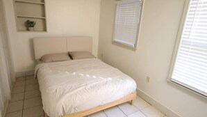 Un dormitorio con cama, una ventana con persianas, un estante con planta y piso de baldosas.