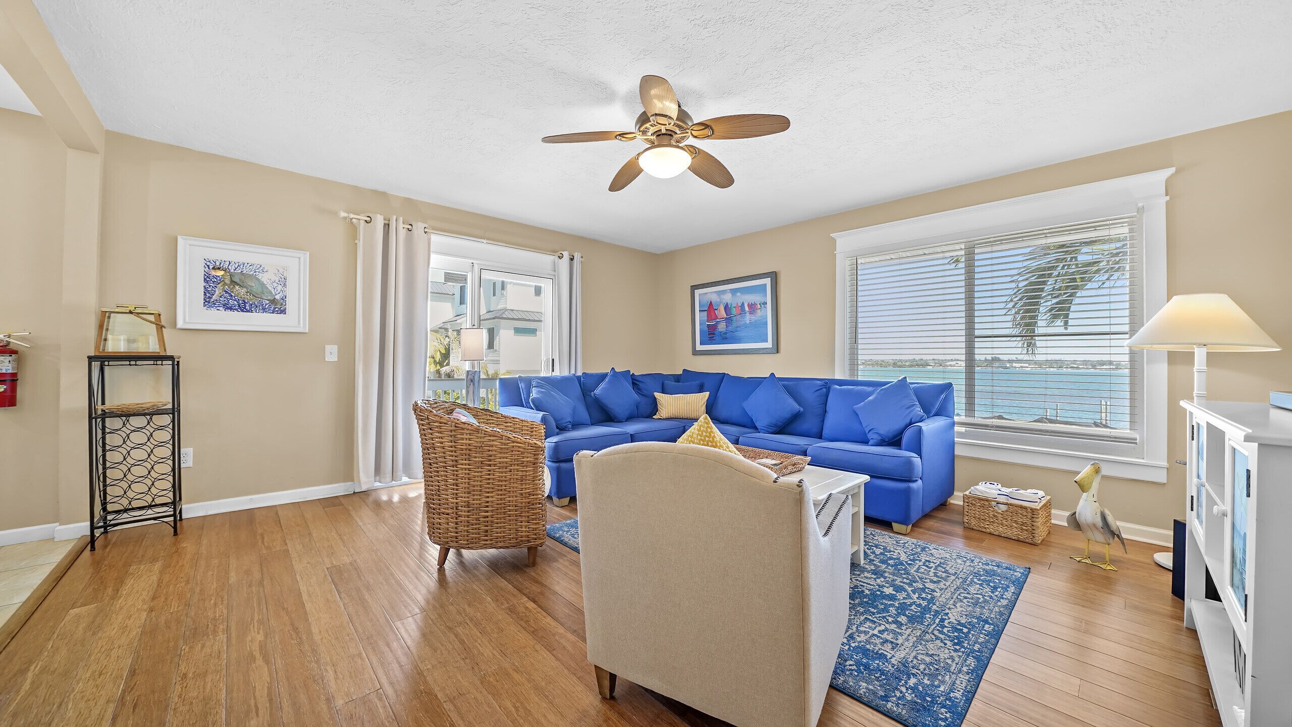 A living room with a blue sofa, a ceiling fan, and a view of the sea through large windows.