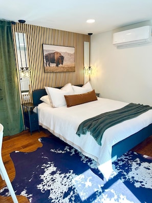 A bedroom with a bed, a nightstand, a mirror, an air conditioner, and a painting of a bison.