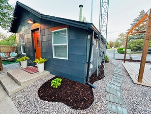 A small house with a dark exterior, a front door, and a window with blinds. There are potted plants in front of the house and a paved area with stepping stones.