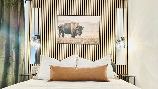 A hotel room with a large bed, striped wall paneling, a mounted picture of a bison, and two wall-mounted lamps. - The Bison Cottage bungalow, a private boutique retreat in Cheyenne. (Cheyenne)