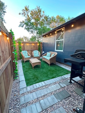 A backyard with a wooden fence, a patio with wicker furniture, a grill, and string lights.