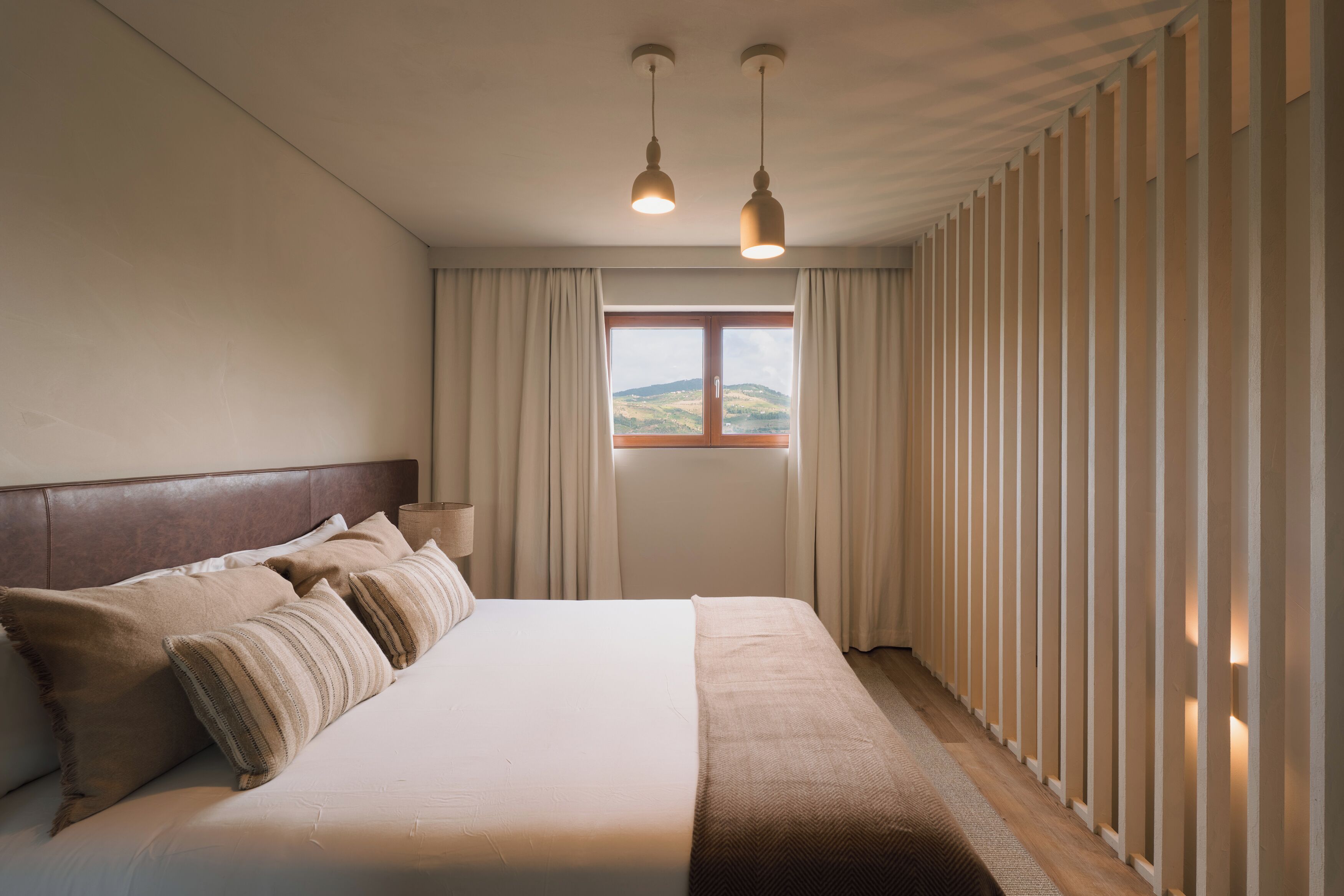 Une chambre à coucher avec un grand lit, deux lampes suspendues, une fenêtre avec des rideaux et une vue sur l’extérieur.