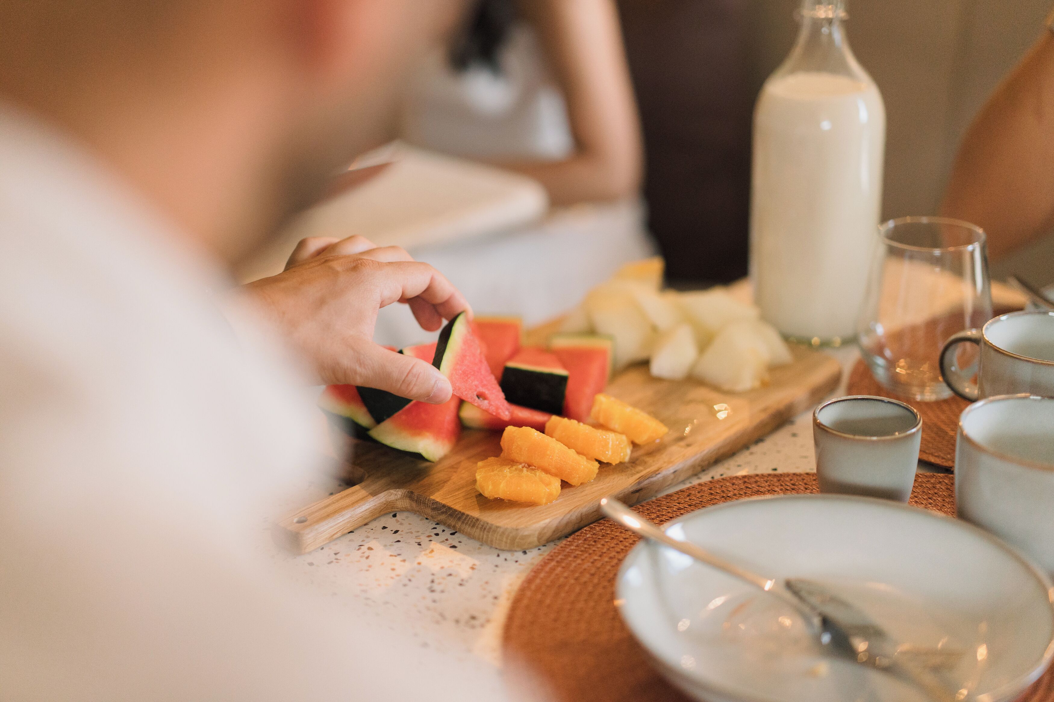 A dining table with a wooden board holding sliced fruits, a bottle of milk, and a glass of milk.