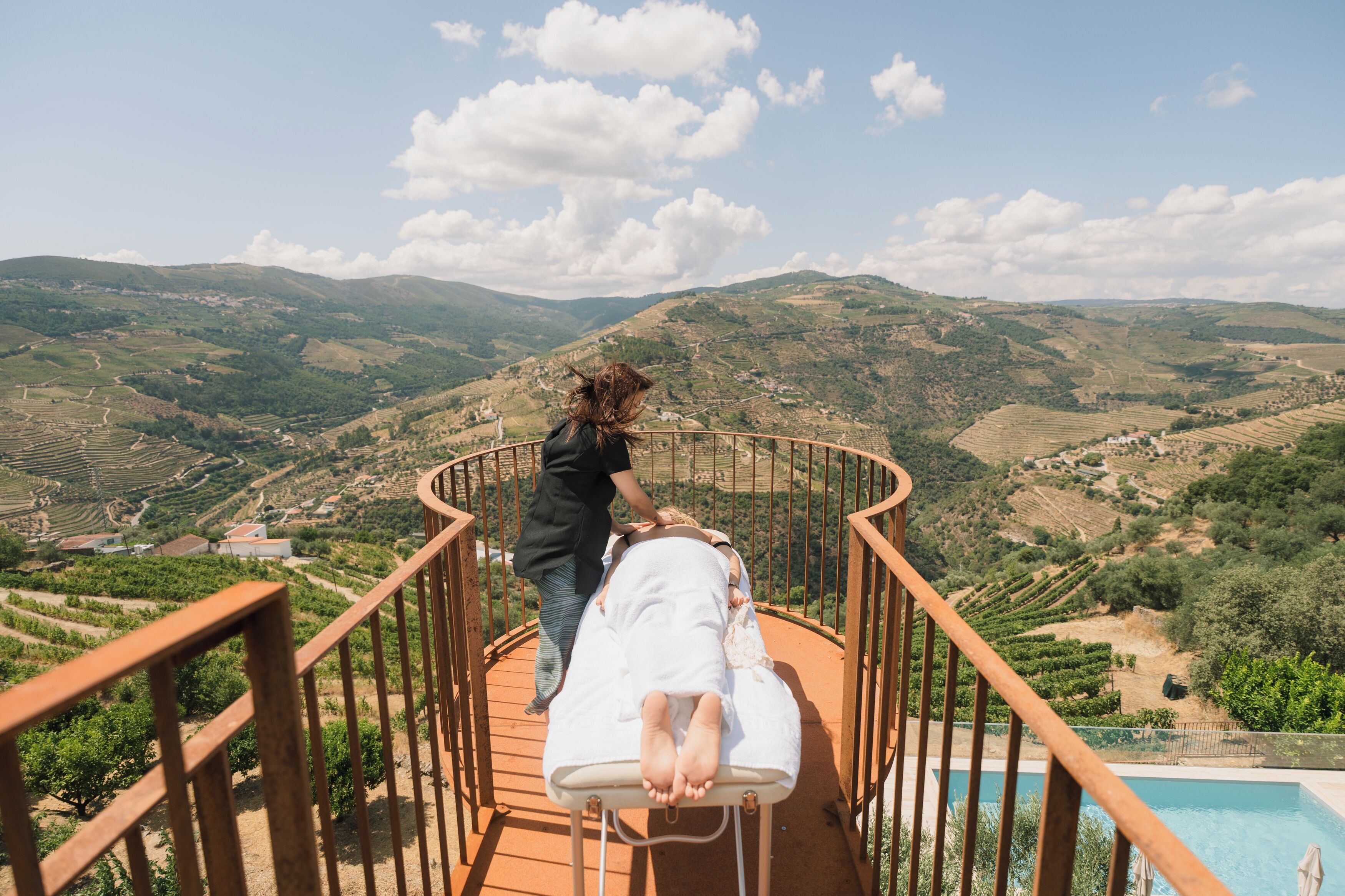 Une personne vêtue d’une robe blanche se tient sur une terrasse en bois, près d’une piscine, et surplombe une vallée où s’étendent des champs en terrasses et des collines.