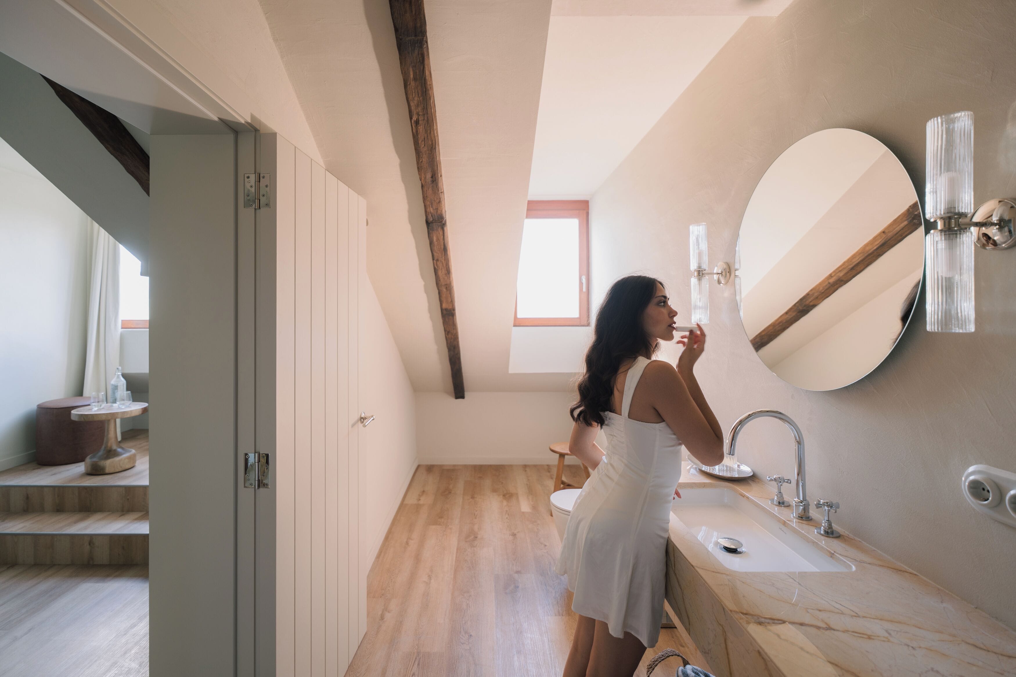A woman in a white dress standing in a bathroom with a large mirror, a sink, and a wooden floor.